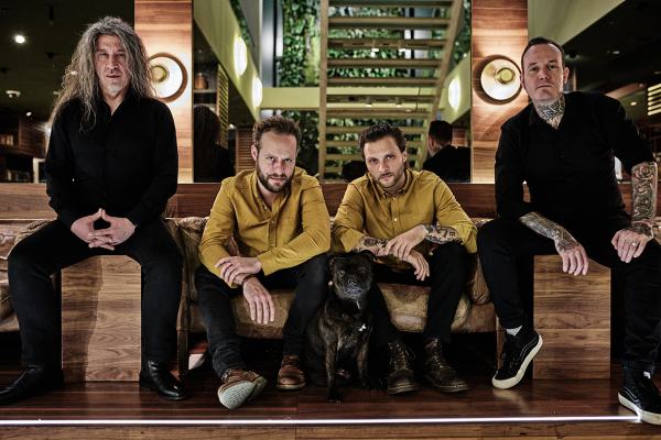 Bandfoto Flying Guillotines: Fünf Personen sitzen und stehen in einer modernen Lobby mit Holztreppe und Pflanzen im Hintergrund. In der Mitte liegt ein schwarzer Hund auf dem Boden. Drei Personen tragen auffällige gelbe Jacken, zwei sind schwarz gekleidet